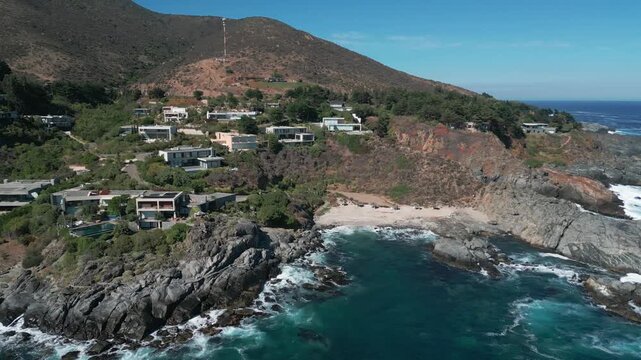 Panoramic shot over exclusive clifftop villas with secluded sandy cove, rugged rocky headland and dry scrubland hill rising behind. Punta Pite, Papudo, Chile