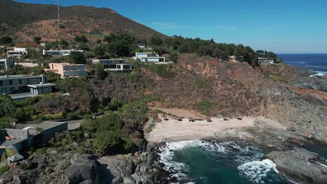 Retreating reveal over secluded sandy cove with crashing waves exposing exclusive modern villas integrated into dramatic rocky clifftop above. Punta Pite, Papudo, Chile