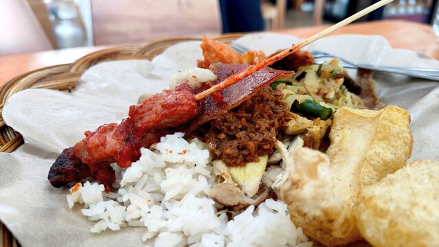 Close up of Balinese nasi babi guling with rice, pork satay, crispy pork skin, pork meat and vegetables served on paper in a woven basket.