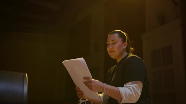 Focused performer rehearsing lines on dimly lit stage, reading script under warm spotlight in quiet auditorium during solitary theatrical practice session