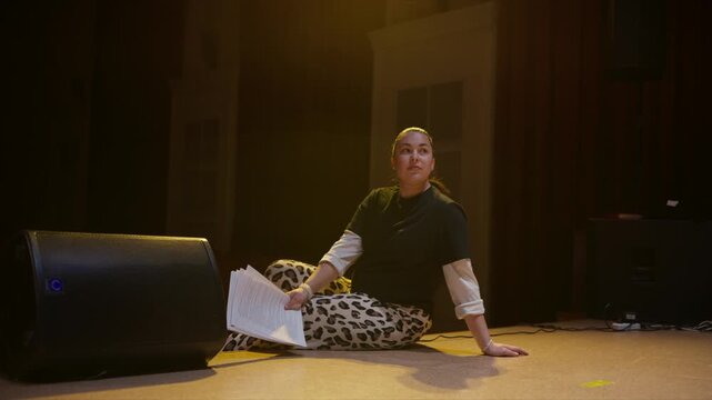 Woman seated on theater stage rehearsing script under warm spotlight, smiling and focused during quiet performance preparation in an empty auditorium setting