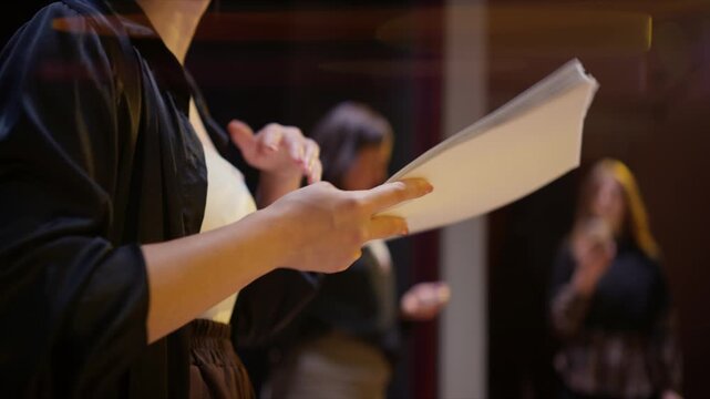 Rehearsal scene with actors reading scripts under dramatic stage lighting, focusing on hands holding pages and blurred performers in a dim theater setting