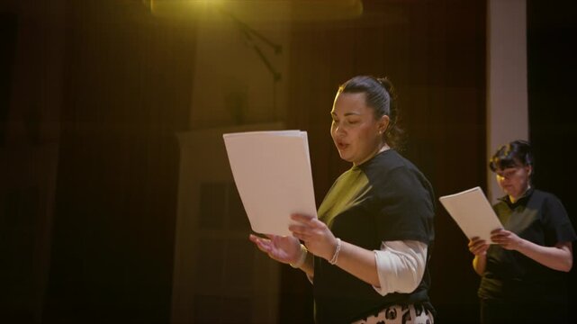 Actors rehearsing lines on a dimly lit stage, holding scripts and phones while practicing dialogue and performance cues in an intimate theater setting