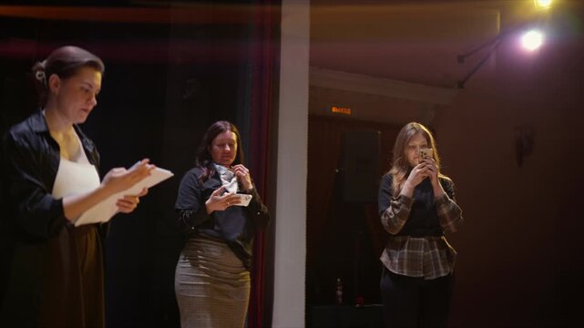 Three women on a dimly lit stage rehearsing with scripts and smartphones, blending traditional performance with modern technology in a dramatic theatrical setting