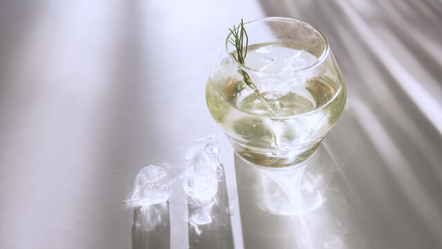 Pale cocktail glass with rosemary garnish, ice and bright caustic reflections. Concept of premium bar menu, cocktail advertising, hospitality design and elegant beverage background.