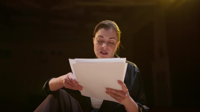 Young performer sitting alone on dimly lit stage reading script pages with focused concentration, rehearsing lines under warm spotlight in empty theatrical auditorium