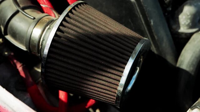 Detailed shot of aftermarket air intake system component under vehicle hood, featuring cylindrical air filter with dark pleats and shiny metallic band, surrounded by other engine parts and bright red 