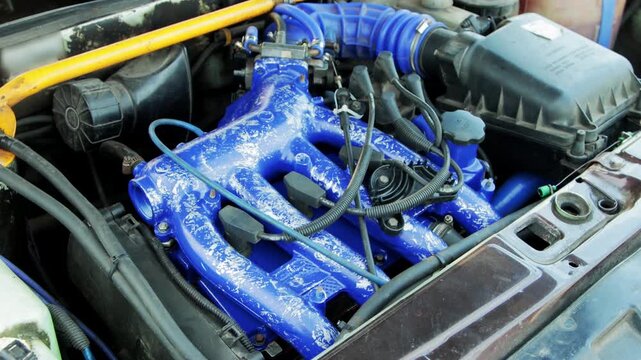 Automobile engine compartment close-up. Network of components. Bright blue intake manifold, textured finish. Hoses, wires, fluid reservoirs, yellow strut bar. Complex mechanical assembly. Automotive p