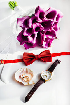 Wedding accessories with purple calla lilies, red bow tie, gold rings and watch on white background