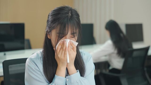 Tired businesswoman office worker female blowing her nose into a paper tissue, suffering symptoms while working on laptop at office desk, sneezing in office, showing the spread of germs and ill health