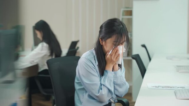 Tired businesswoman office worker female blowing her nose into a paper tissue, suffering symptoms while working on laptop at office desk, indicating fatigue and potential viral infection cold symptoms