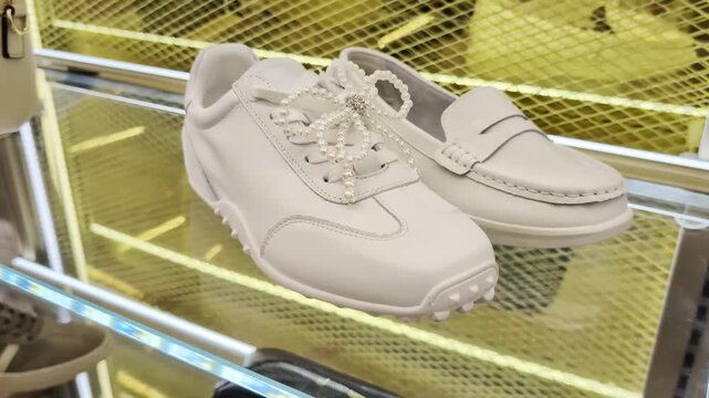 Two white shoes, one sneaker with decorative pearl bow, one classic loafer, displayed on transparent glass shelf. Background features golden grid pattern. Footwear showcase presents clean design, luxu