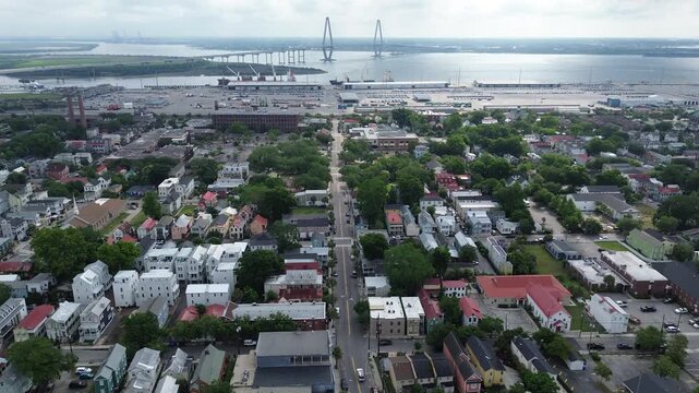 Harbor bridge corridor frames Meeting St residential grids extending toward industrial waterfront parcels, Ravenel Bridge Cooper River. Mixed roofing, tree pockets, ports, rails, Charleston, SC