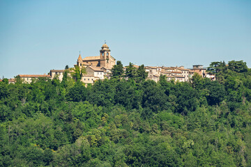 Fototapeta premium Mondaino village on its hill in Montefeltro, Italy, with the church of San Michele Arcangelo rising above green forest under clear sky