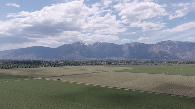 Orbiting aerial view of lush green farmland stretching towards the snow-capped Sierra Nevada mountains under a partly cloudy sky, showing the vast rural landscape of Minden, Nevada