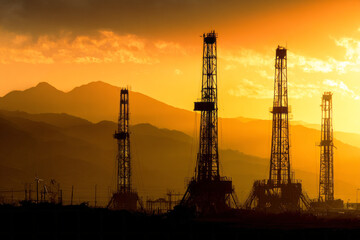 Obraz premium Silhouettes of oil and natural gas drilling rigs stand in front of mountains as the sun sets, creating a colorful sky. The scene shows energy production in a natural setting