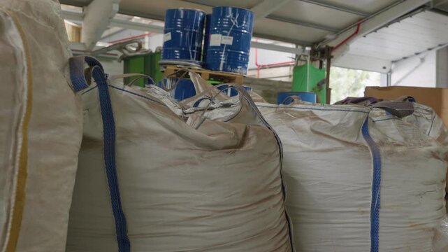 Rows of filled white bulk bags with blue handles and stacked blue chemical drums inside recycling warehouse