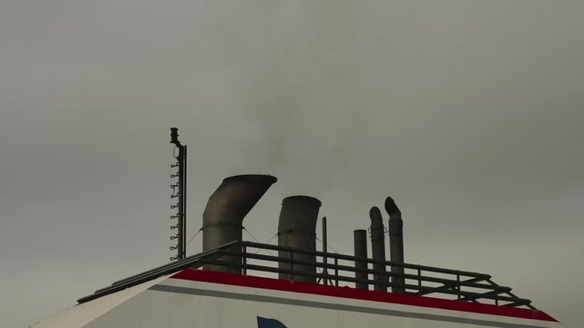 Multiple exhaust funnels and antenna mast rise from ferry upper deck with red stripe under grey sky