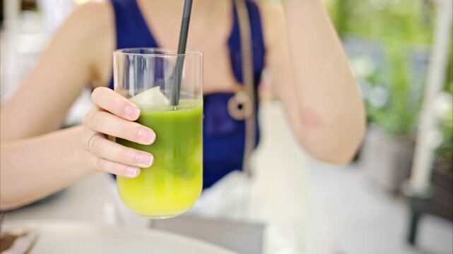 Woman stirs refreshing iced matcha green tea beverage in clear glass sitting at outdoor cafe during summer time.