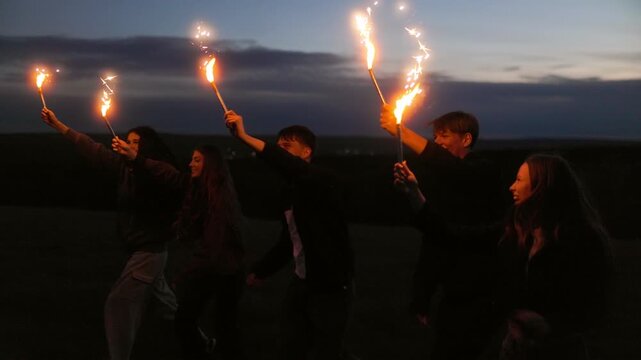 Happy group of multiethnic teenagers running with bright sparklers at night.