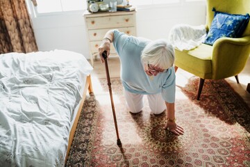 Elderly woman with a cane kneeling on a patterned rug in a cozy bedroom.