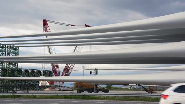 Rows of long white wind turbine blades stored on green steel racks beside a road with a tall crane a speed limit sign and an overcast sky at an industrial storage site