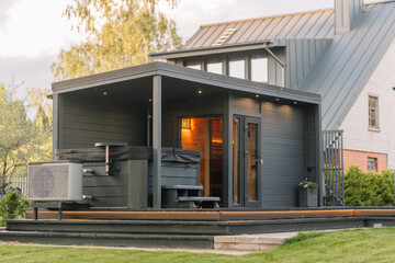 Naklejka premium Modern wooden sauna with hot tub and outdoor deck situated in a residential backyard surrounded by greenery and a contemporary house in the background