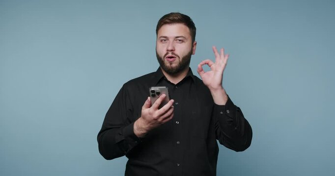 Man speaks and gestures while using phone in studio with blue background during daytime to share information
