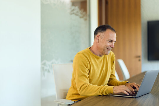Smiling Middle-Aged Man Working Remotely on Laptop in Modern Office Conference Room for Business