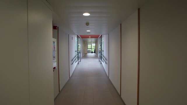 Long empty corridor inside a modern building with handrails on both sides glass partitions ceiling lights and wall mounted fire safety equipment leading to a seating area