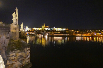 Obraz premium Charles Bridge glows over the Vltava at night, where Gothic towers, stone arches, and golden reflections create one of Prague’s most atmospheric river scenes.
