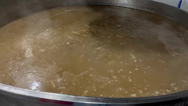 Stock broth boiling in a large industrial cooking kettle during food manufacturing operations. Soup base preparation process using ingredients and seasonings to create consistent flavor for large-scal