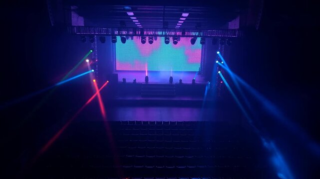 Static wide shot of empty concert stage with colorful moving spotlights and led screen in dark theater auditorium