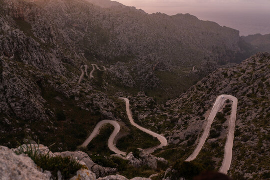 Sa calobra road carves its serpentine path through jagged rocky mountains, offering a challenging yet scenic drive with switchbacks leading to the mediterranean coastline under a soft sunset sky