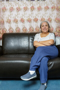 Smiling, makeup-free, haggard adult woman sits cross-legged with her arms resting in her lap, maintaining eye contact with the camera, decorative fabrics are visible on the background wall