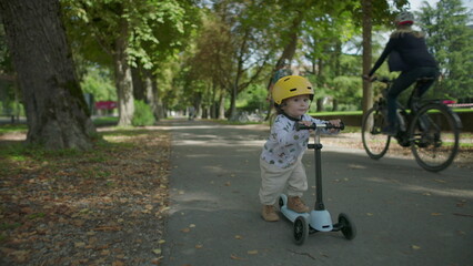 Naklejka premium Small child riding scooter toward camera on park path, wearing protective yellow helmet, concentrating with determination while practicing balance and movement