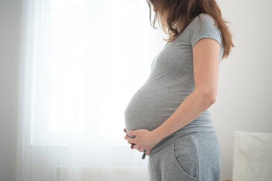 Pregnant woman by window hands on stomach in serene maternity portrait. Expecting mother with tilted head in bright comfortable bedroom. Future parent cradling belly in elegant peaceful home.