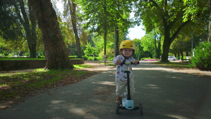 Naklejka premium Small toddler wearing yellow helmet riding scooter along tree-lined park path, moving with curiosity and energy while exploring outdoor environment
