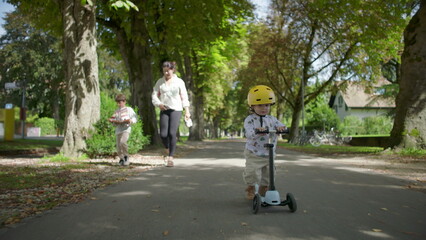 Obraz premium Toddler riding scooter on pathway wearing yellow helmet, mother and older sibling walking behind, family enjoying time together outdoors in peaceful park setting