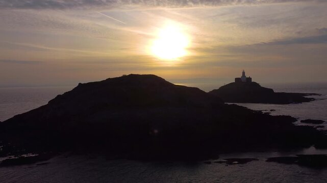 Rising Aerial of Mumbles Lighthouse at Sunrise. Golden Reflection of Sun on Ocean with Dynamic Clouds. Perfect for tourism, travel documentaries. Wales, UK. 4K.