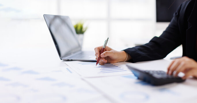 Businesswoman analyzing financial charts and reports, marking figures with a pen while using a calculator and laptop for budgeting, accounting and investment planning in an office