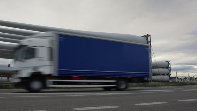 A white car drives in motion blur along a highway past a stack of huge white wind turbine blades resting on steel racks at an industrial storage yard under a grey sky
