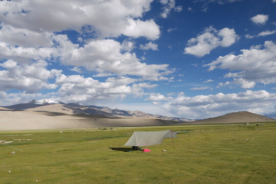 A camping tarp shelter pitched on a vast green alpine meadow against majestic desert mountain ranges under a beautiful cloudy blue sky in Ladakh, Himalayas.