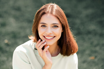 Fototapeta premium Smiling young woman portrait outdoors looking at camera with hand near face, natural light and casual clothing conveying friendly, confident lifestyle and wellness mood