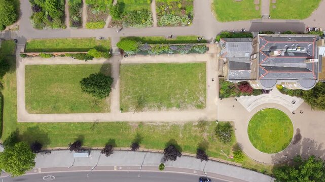 Drone view over Lloyd Park and William Morris House, Walthamstow, London. Top-down forward aerial reveals lawns, paths, circular plaza and nearby road traffic in sunshine.