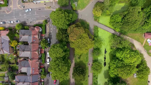 Victorian terraced houses, street traffic and rooftops on the left, with green lawns, trees and walking paths in Lloyd Park, Walthamstow, London, UK to the right, showing contrast of urban and nature.