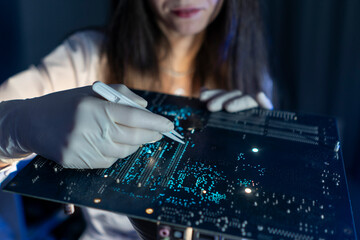 A person wearing gloves uses a pen to mark a circuit board in a lab. The setting is dark with blue...
