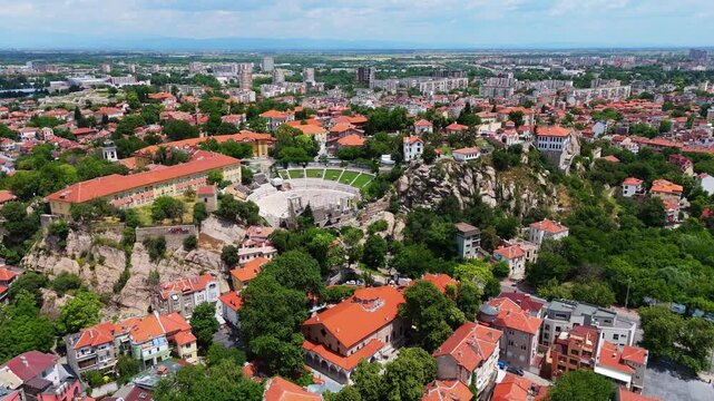 Aerial drone tracking shot of the ancient Roman Theatre of Philippopolis next to a modern road tunnel, revealing traditional houses on the rocky hillside of Plovdiv, Bulgaria
