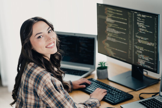 Smiling female programmer coding at a dual-monitor desk with laptop in a bright home office, young software developer working on web development and technology