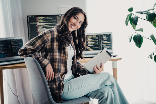Smiling woman programmer working from home office with tablet and multiple laptops showing code, casual remote developer in modern workspace